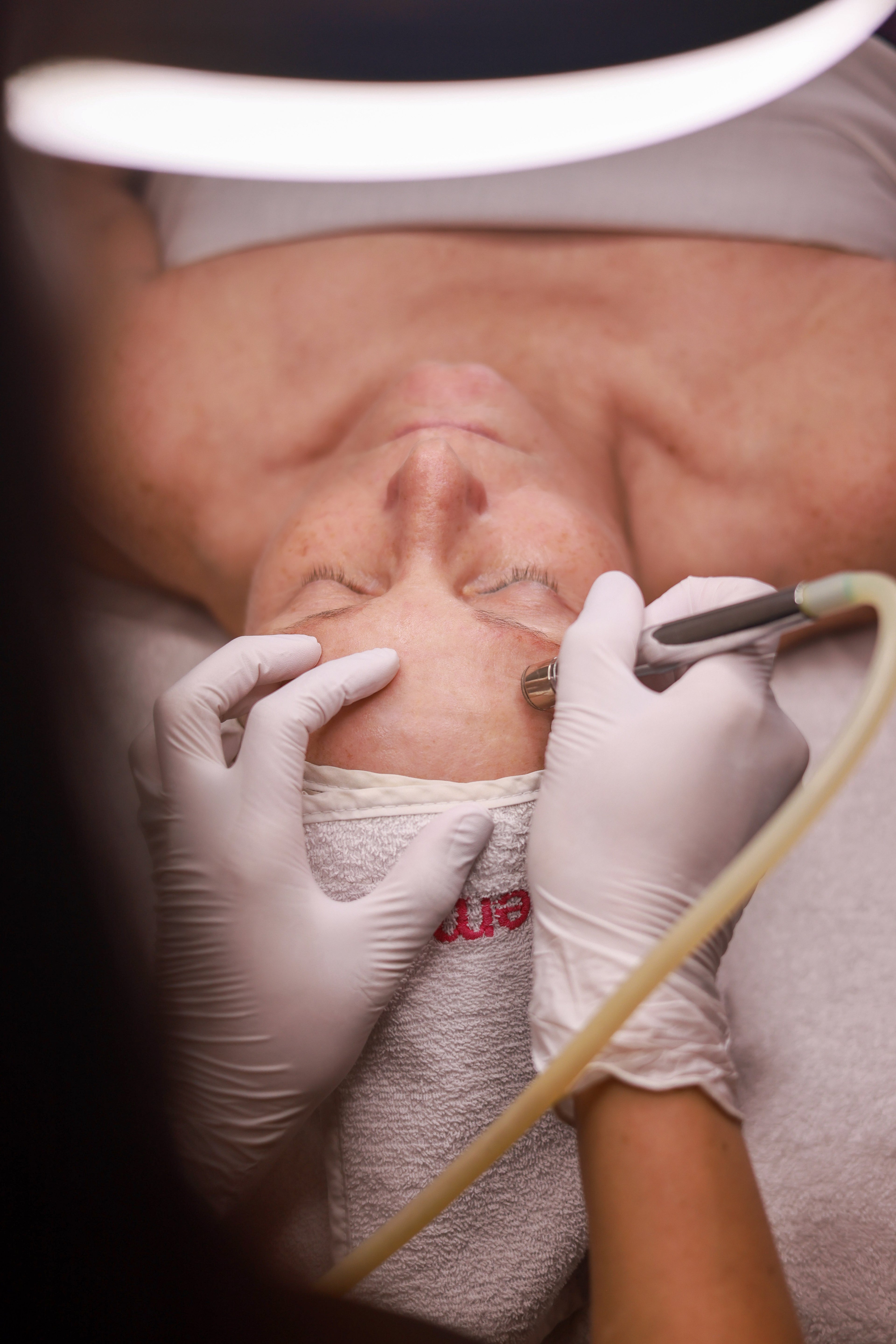 A person receives a facial treatment with a microblading tool by a technician wearing gloves. The setting is calm and professional.