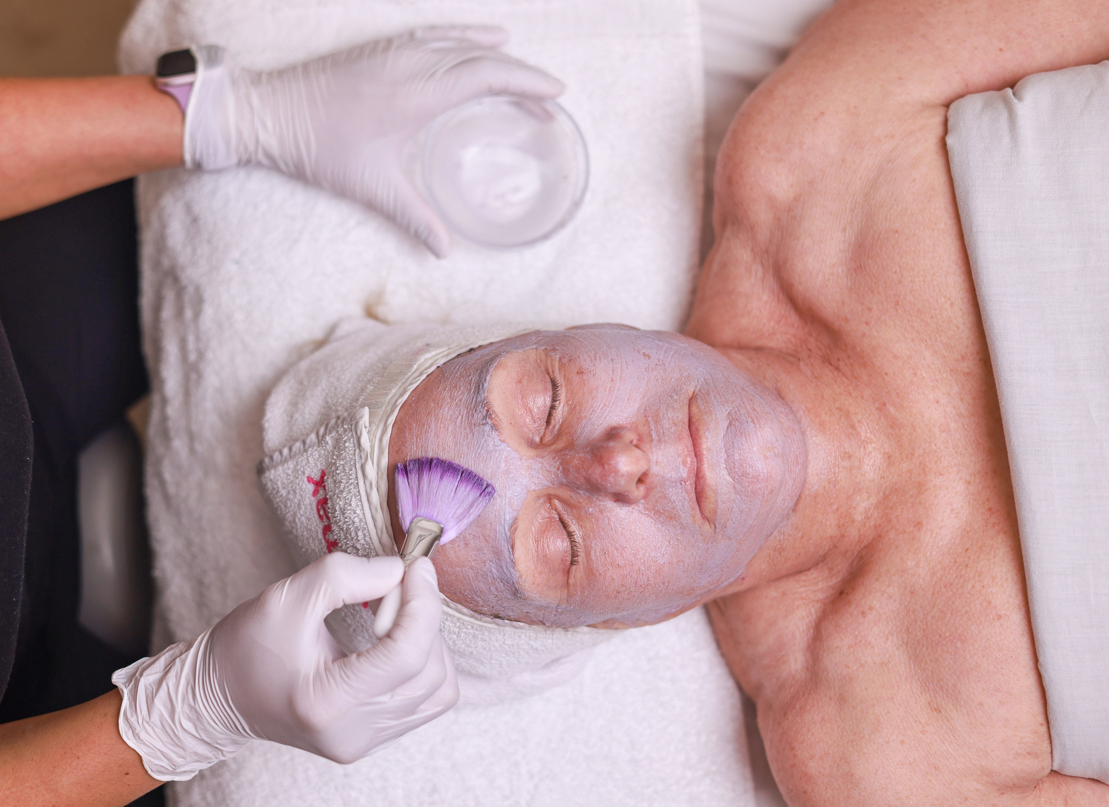 A person receives a soothing purple facial mask in a spa setting, with eyes closed. A beautician applies the mask using a brush, wearing gloves.
