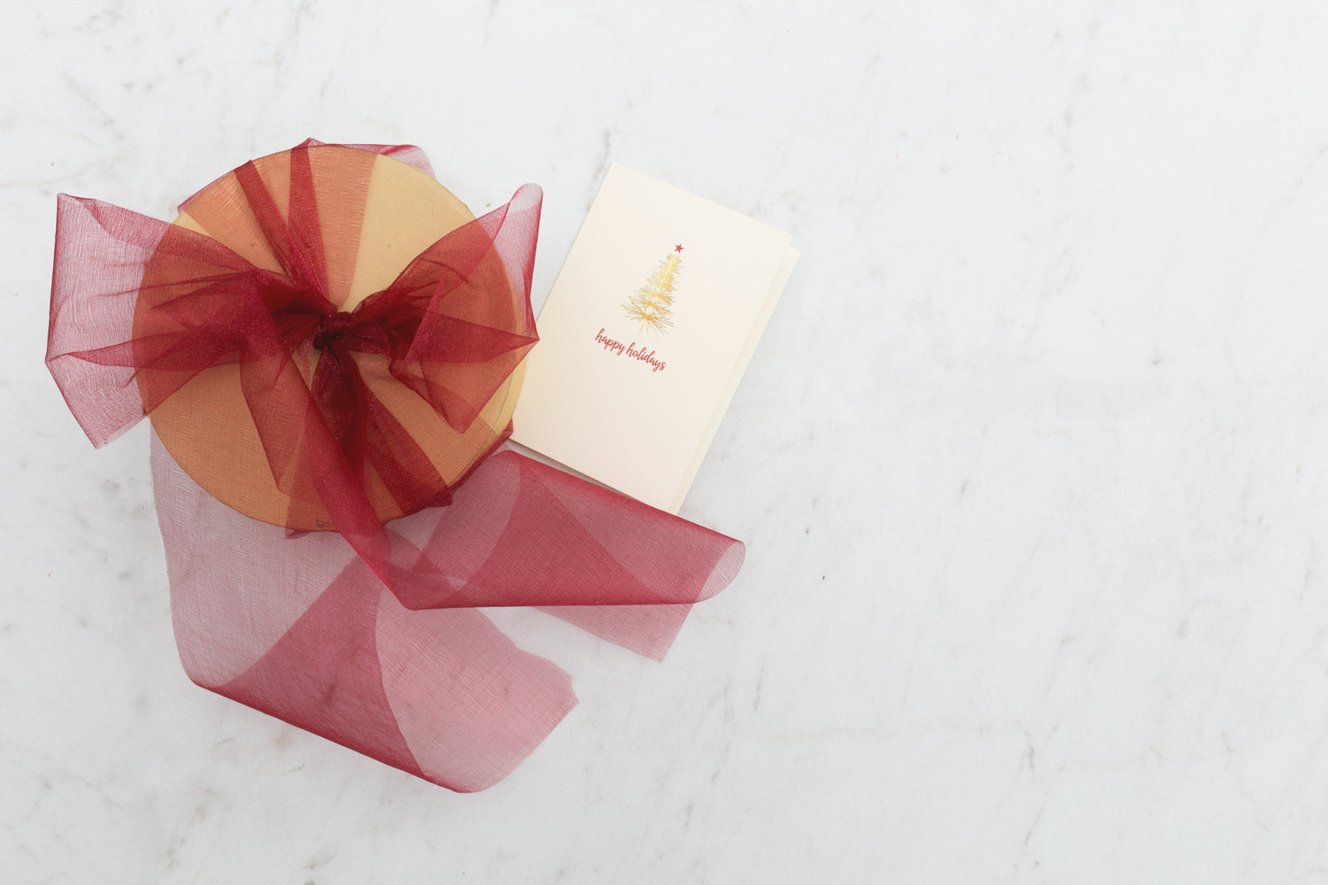 A round gift box wrapped with a deep red bow sits beside a holiday card with a gold tree design, on a clean white marble surface.